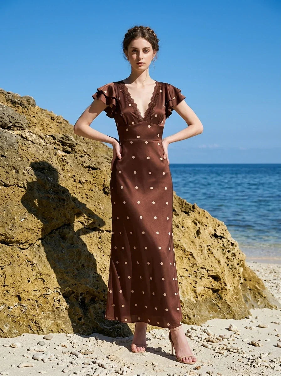 Woman wearing a brown polka dot midi dress with ruffled sleeves and a v-neckline, posing with hands on hips on a beach.
