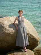 Woman wearing a black and white gingham halter maxi dress leaning against a large rock on a beach