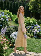 Woman wearing a sleeveless yellow maxi dress with a floral jacquard pattern walking in a garden with hydrangeas and other flowers.
