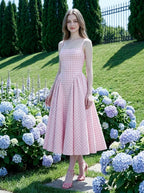 A woman models a pink and white gingham midi dress with a square neckline and a full, pleated skirt, standing amongst blue hydrangeas in a garden.