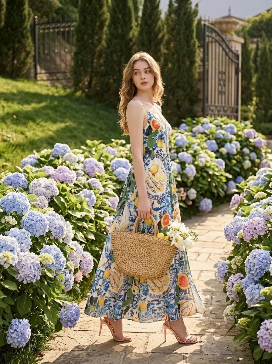 Woman wearing a long, sleeveless maxi dress with a colorful fruit and floral print, holding a straw bag and walking in a garden.