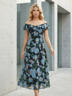 Woman standing in an urban setting wearing a black off-the-shoulder midi dress with a blue floral print