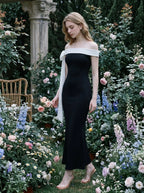Woman standing in a black off-the-shoulder mermaid gown with a white sash and bow detail amidst a floral garden.