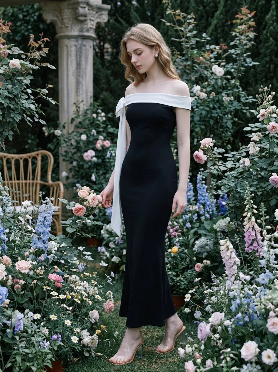 Woman standing in a black off-the-shoulder mermaid gown with a white sash and bow detail amidst a floral garden.