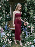Woman standing in a garden wearing a burgundy slip dress with lace waist detailing and touching flowers.