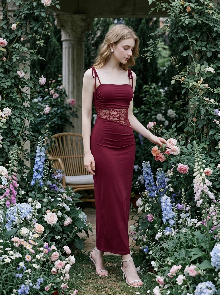 Woman standing in a garden wearing a burgundy slip dress with lace waist detailing and touching flowers.