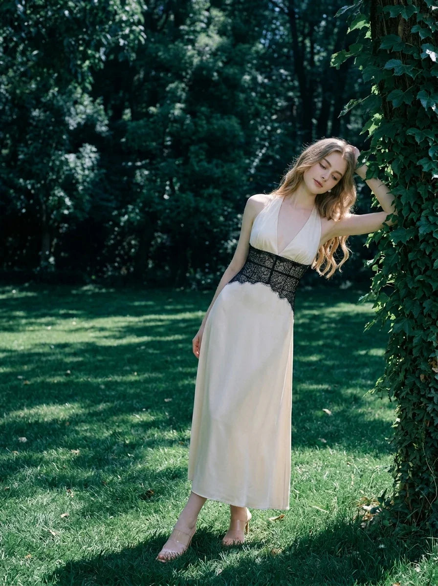 Woman in a cream satin maxi dress with black lace waist detail, leaning against a tree in a garden.