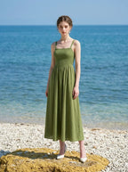 Woman wearing a green linen midi dress with a smocked bodice and square neckline, standing on a rocky shore with the ocean behind her.