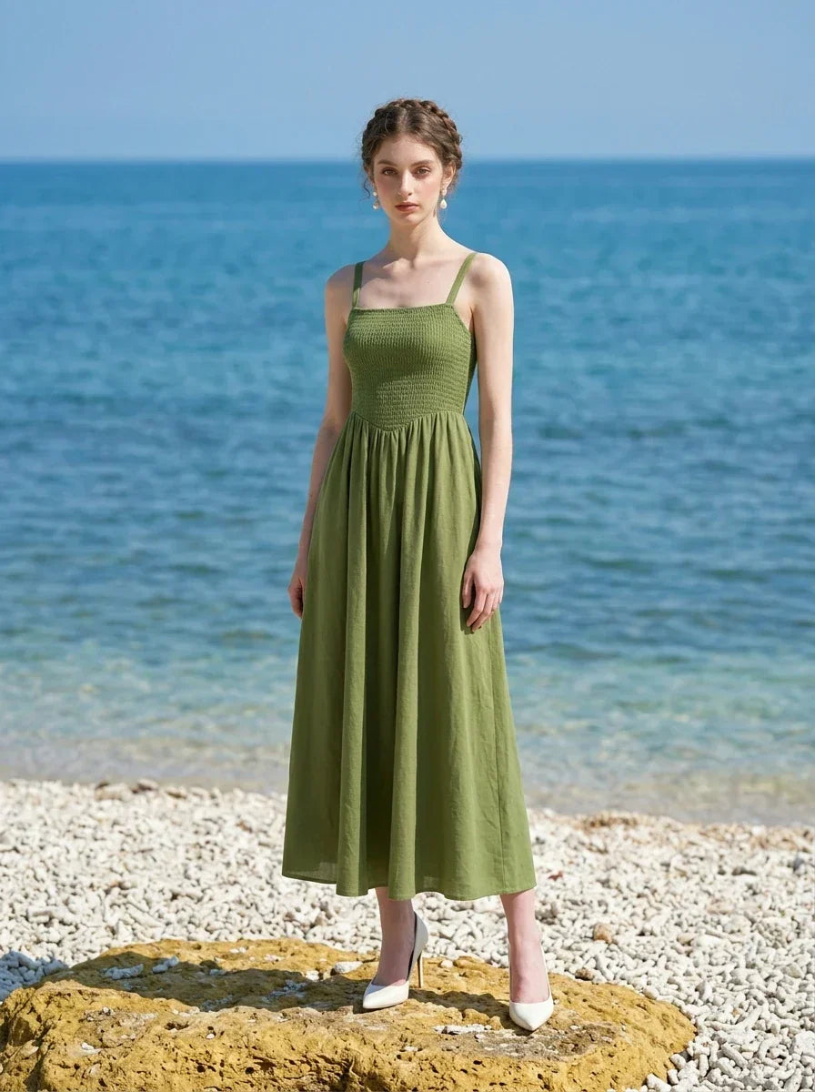 Woman wearing a green linen midi dress with a smocked bodice and square neckline, standing on a rocky shore with the ocean behind her.