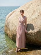 Woman in a rose gold shimmering metallic A-line V-neck sleeveless floor-length formal gown, posed by a rock near the ocean