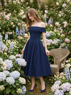 Woman wearing a navy blue off-the-shoulder midi dress with a pleated skirt, standing in a garden with flowers and a wicker chair.
