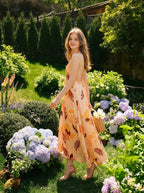 Woman walking in a garden wearing a long, floral print, halter neck dress with an open back, carrying a straw basket.