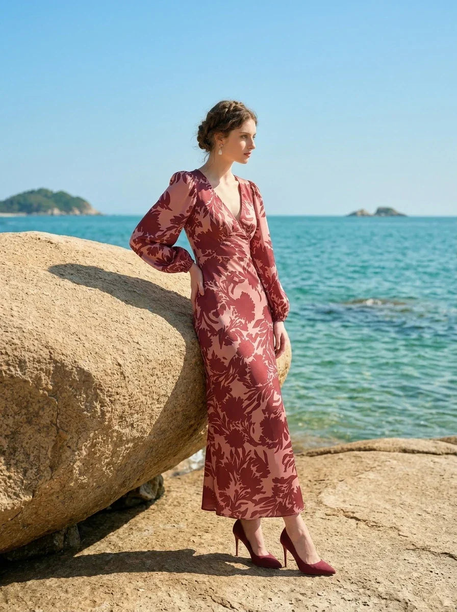 woman wearing a pink and red floral print satin maxi wrap dress with long bishop sleeves and a v-neck, leaning against a large rock on a beach with the ocean in the background