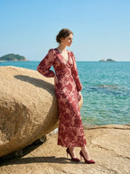 woman wearing a pink and red floral print satin maxi wrap dress with long bishop sleeves and a v-neck, leaning against a large rock on a beach with the ocean in the background