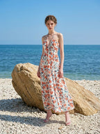 Woman wearing a white floral print maxi dress with a halter neckline and tiered skirt, standing on a sandy beach next to a large rock with the ocean in the background.