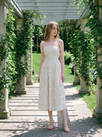 Woman wearing a white floral print midi dress with spaghetti straps and a square neckline, standing on a stone pathway under a pergola.