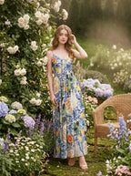 Woman wearing a long, sleeveless maxi dress with a colorful fruit and floral print, posing in a garden with roses and hydrangeas.