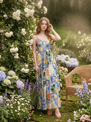 Woman wearing a long, sleeveless maxi dress with a colorful fruit and floral print, posing in a garden with roses and hydrangeas.