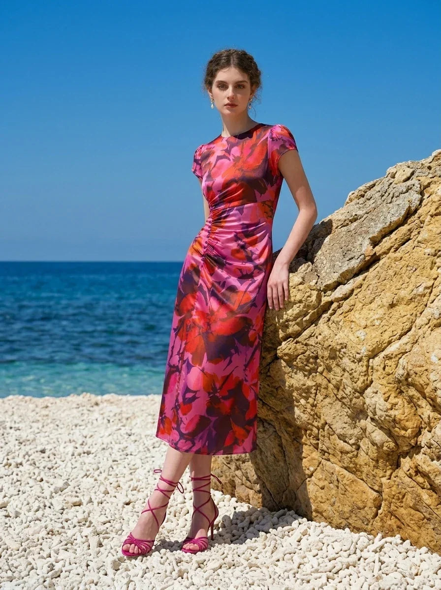Woman wearing a pink and red floral print satin midi dress with short sleeves and ruched detailing, leaning against a rock on a beach with the ocean in the background.