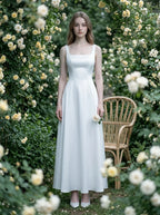 Woman wearing a white satin sleeveless A-line wedding dress with a square neckline, standing in a garden with yellow roses