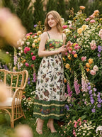 Woman posing in a garden with flowers, wearing a floral and bird print midi dress with spaghetti straps and a square neckline.