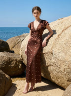 Woman wearing a brown polka dot midi dress with ruffled sleeves and a v-neckline, leaning against a large rock on a beach.
