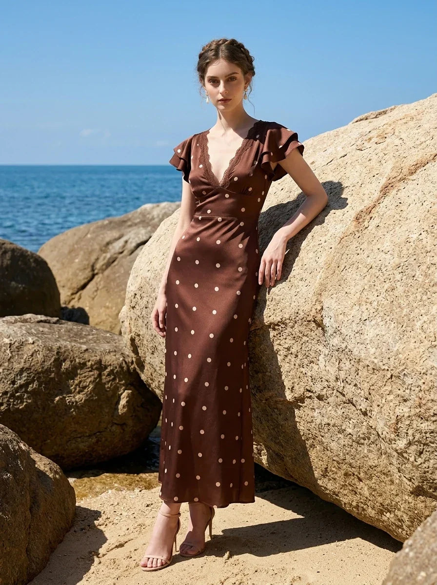 Woman wearing a brown polka dot midi dress with ruffled sleeves and a v-neckline, leaning against a large rock on a beach.