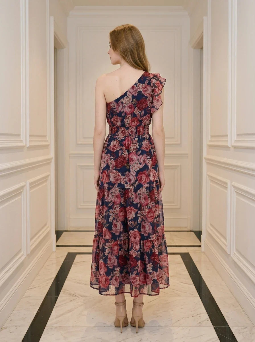 Back view of a woman wearing a navy floral print one-shoulder dress with a tiered skirt and ruffle detail, standing in a hallway.