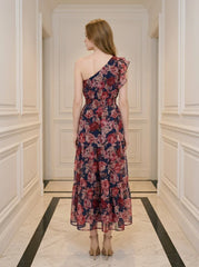Back view of a woman wearing a navy floral print one-shoulder dress with a tiered skirt and ruffle detail, standing in a hallway.