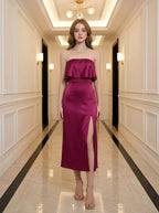 Woman wearing a strapless magenta satin midi dress with a high slit, standing in a hallway.