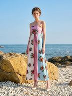 woman in a pink, blue, and white floral print maxi dress with spaghetti straps, standing on a rocky beach with the ocean in the background