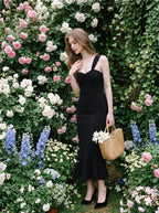 Woman in a black asymmetrical one-shoulder midi dress with a ruffled hem, holding a straw bag, standing among roses and blue flowers.