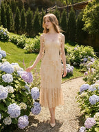 Woman wearing a cream-colored sleeveless midi dress with floral embellishments and a ruffled hem, walking through a garden with hydrangeas.