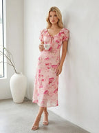 Woman in a pink floral print midi dress holding a teacup while leaning against a wall.