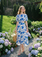 Woman wearing a white and blue floral print midi dress with cut-out waist and puff sleeves, holding a straw bag in a garden.
