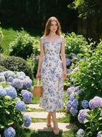 Woman wearing a floral print midi dress with tie shoulder straps and a high slit, walking in a garden with hydrangeas.