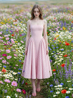 A woman models a pink and white gingham midi dress with a square neckline and a full, pleated skirt, walking through a field of wildflowers.