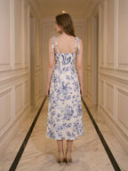 Back view of a woman wearing a floral print midi dress with tie shoulder straps and a high slit, standing in a hallway.