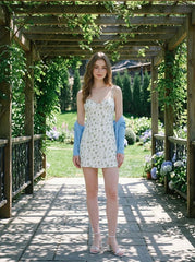 Woman wearing a white floral print mini dress with spaghetti straps and a sweetheart neckline, with a blue cardigan draped over her shoulders, standing in a garden arbor.