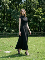 Woman in a black mock neck cap sleeve A-line dress walking in a grassy park with trees in the background.