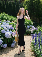 Woman in a black asymmetrical one-shoulder midi dress with a ruffled hem, carrying a straw bag, walking in a garden with hydrangeas.