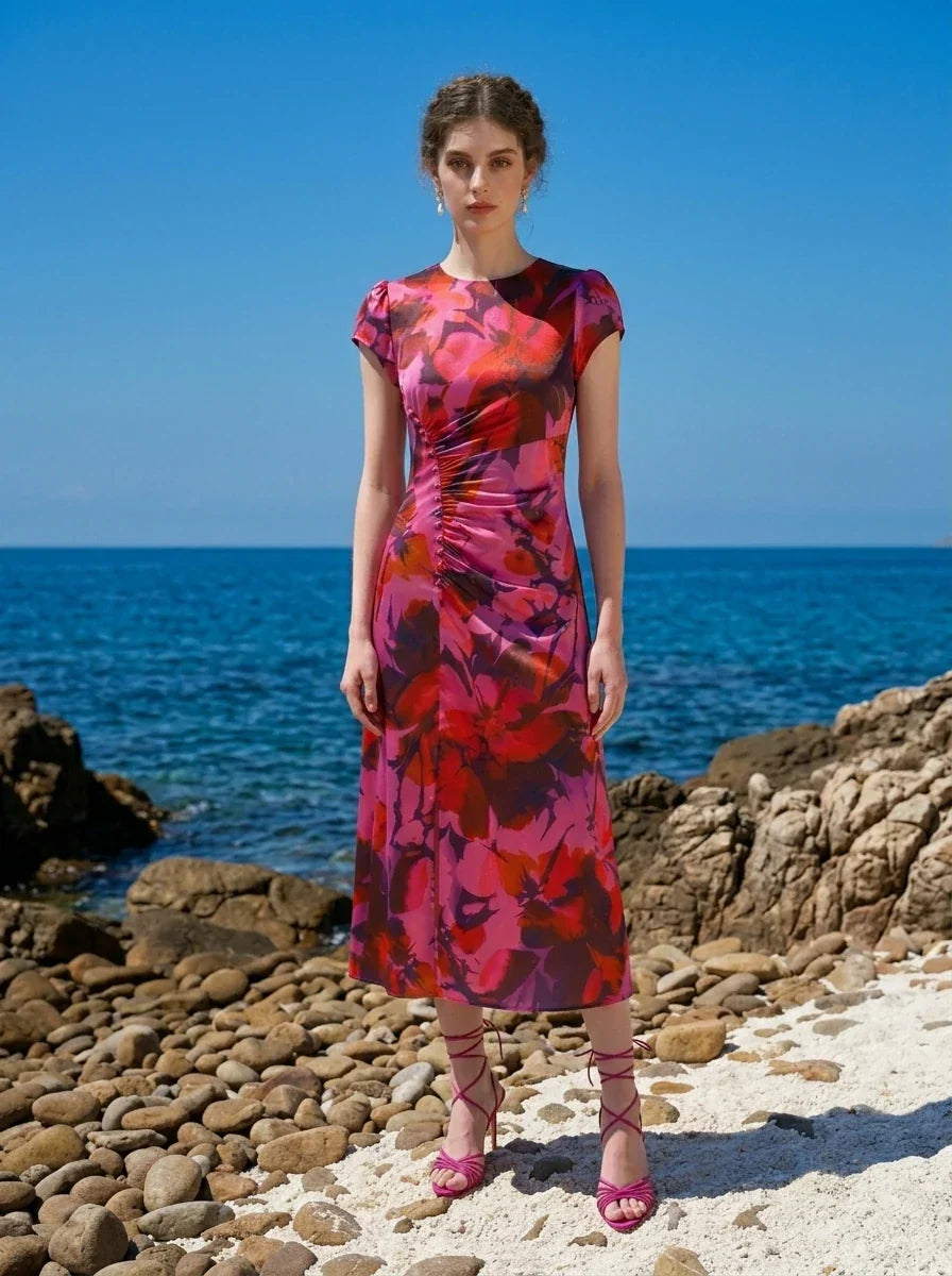 Woman wearing a pink and red floral print satin midi dress with short sleeves and ruched detailing, standing on a rocky beach with the ocean in the background.