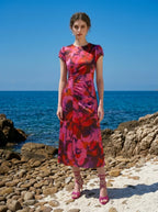 Woman wearing a pink and red floral print satin midi dress with short sleeves and ruched detailing, standing on a rocky beach with the ocean in the background.