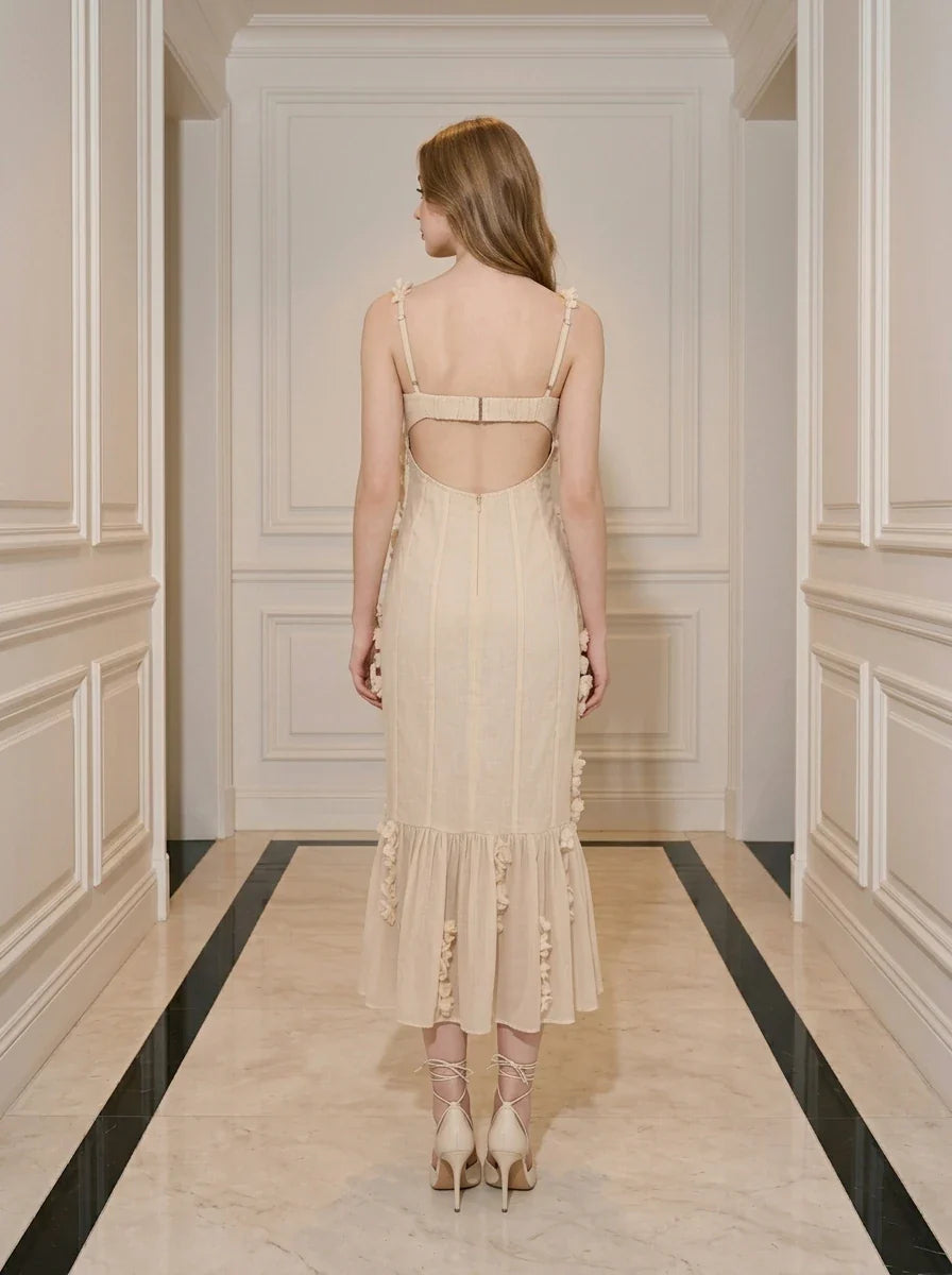 Back view of a woman wearing a cream-colored sleeveless midi dress with floral embellishments and a ruffled hem, standing in a hallway.