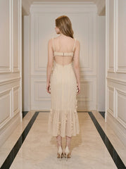 Back view of a woman wearing a cream-colored sleeveless midi dress with floral embellishments and a ruffled hem, standing in a hallway.