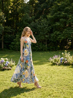 Woman wearing a long, sleeveless maxi dress with a colorful fruit and floral print, walking in a grassy garden area.