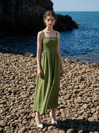 Woman wearing a green linen midi dress with a smocked bodice and square neckline, posing on a pebble beach with the ocean in the background.