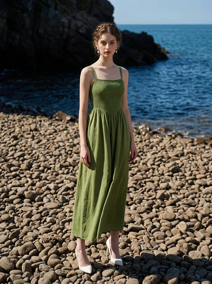 Woman wearing a green linen midi dress with a smocked bodice and square neckline, posing on a pebble beach with the ocean in the background.