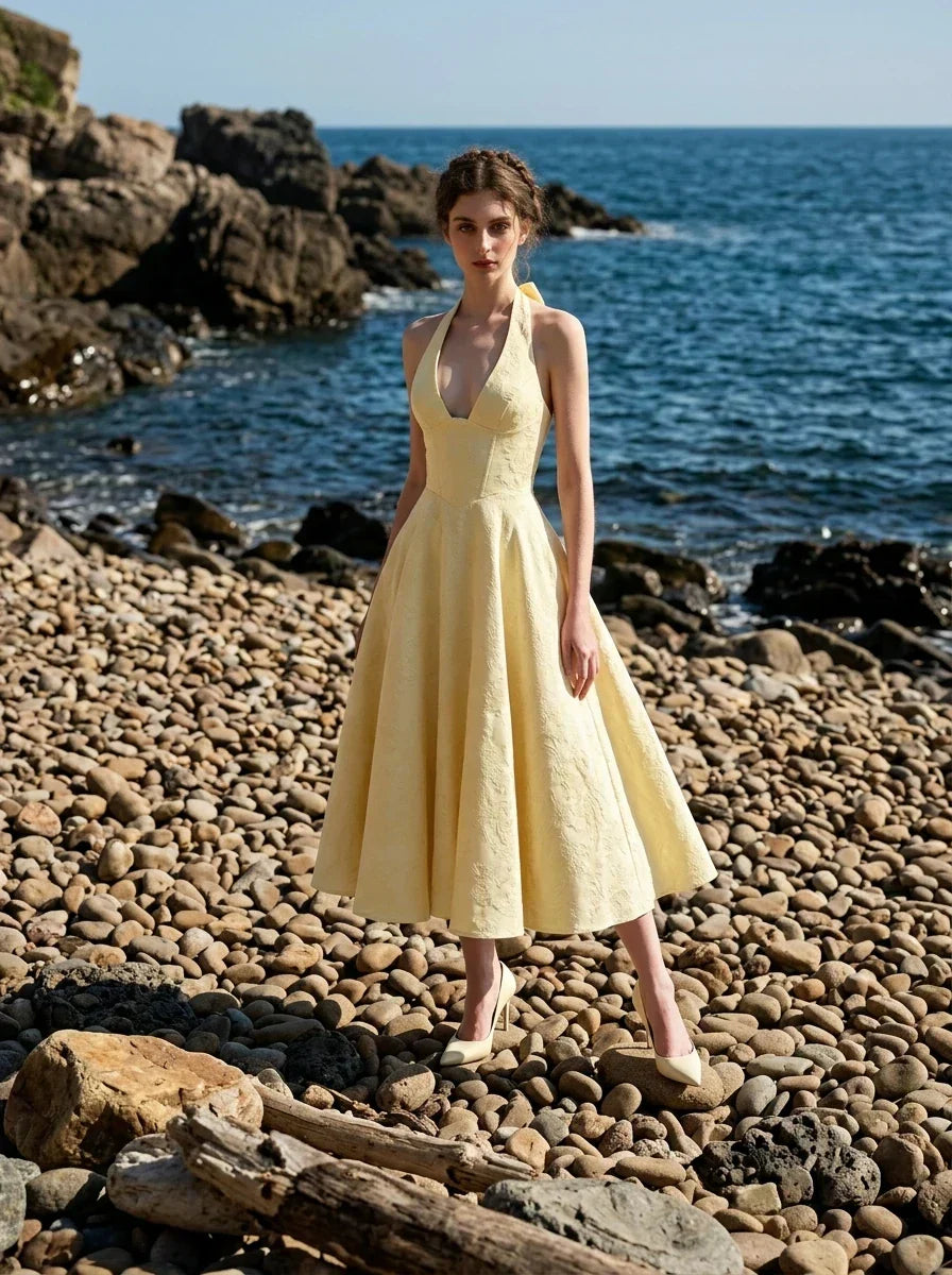 Woman wearing a yellow halter neck midi dress with a full skirt, posing on a rocky beach with the ocean in the background.