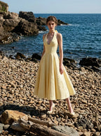 Woman wearing a yellow halter neck midi dress with a full skirt, posing on a rocky beach with the ocean in the background.